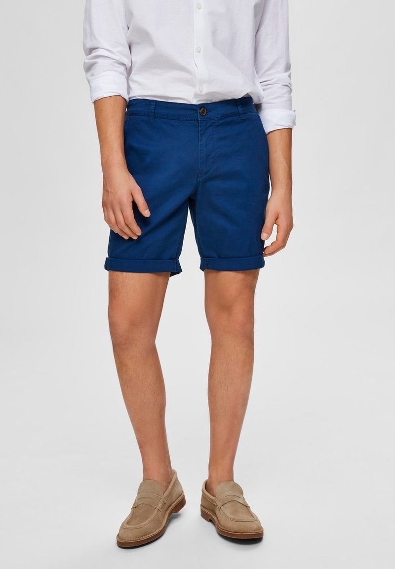 Blå bomullsshorts med rullade fållar, med en framknappning och sidofickor, kombinerat med en vit skjorta och beiga loafers.