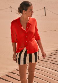 Rotes leichtes Hemd mit Knopfleiste und Kragen, kombiniert mit schwarz-weiß gestreiften Shorts mit vertikalen Streifen. Glatter Stoff, entspannte Passform.