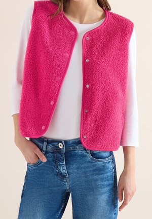 Femme portant un gilet sans manches en polaire fuchsia rose sur une chemise blanche à manches longues et un jean bleu, avec une main dans la poche.