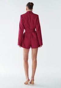 Bordový na mieru šitý overal s dlhými rukávmi, vypasovaným pásom a šortkami. Má vysoký golier a detail so zipsom na chrbte. Hladká textúra.