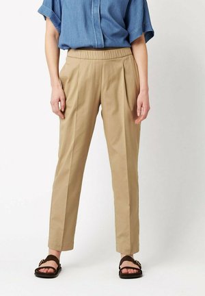 Person wearing beige tapered pants with pleats, an elastic waistband, a blue rolled-sleeve shirt, and dark sandals standing against a plain background.
