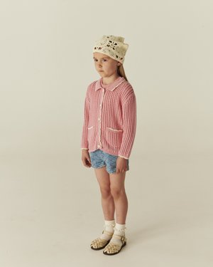 Kind steht, trägt eine cremefarbene gehäkelte Mütze, rosa Strickjacke, blaue gesteppte Shorts, cremefarbene Socken und beige gewebte Sandalen.