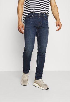 Homme portant un jean slim bleu foncé, des baskets beiges et une chemise à rayures noires et blanches, debout devant un fond uni.