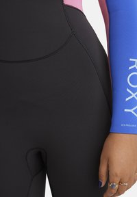 Close-up van een persoon die een zwart wetsuit draagt met een helderblauwe mouw met de tekst "ROXY", en een hand toont met blauwe nagellak en ringen.