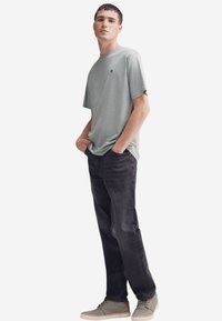 T-shirt in cotone verde chiaro a maniche corte, abbinata a jeans in denim grigi scuri. Il modello indossa sneakers grigie con suole bianche, che mostrano uno stile casual.