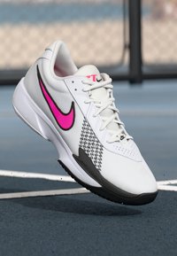 Hvid sportssko med en lyserød Nike swoosh, tekstureret overdel, sorte detaljer og et mønster på siden. Har snørebåndsdesign og polstret sål.