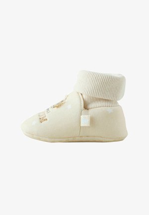 Beige Babyschuhe aus weichem Baumwollmaterial, mit geripptem Bündchen und strukturierter Sohle. Mit gesticktem Design und kleinem Etikett an der Seite.
