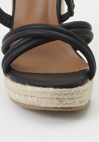 Sandalo in pelle nera con cinturini, dotato di una spessa piattaforma in juta intrecciata e di una suola in morbida pelle marrone. Design accattivante delle cinghie incrociate nella parte anteriore.