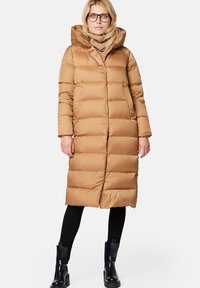 Brązowa kurtka puchowa z kapturem, z poziomymi przeszyciami, bocznymi kieszeniami i długością do połowy łydki. Nosi ją z jasnobrązowym golfem.
