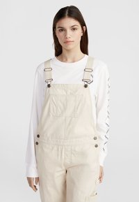 Beige cotton overalls with adjustable straps, front pocket, and side buttons, worn over a white long-sleeve shirt with patterned sleeve detailing.