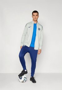 Gray zip-up jacket with blue accents, blue T-shirt, and navy joggers. Model stands on a soccer ball, wearing black athletic shoes.