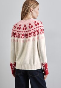Strickpullover mit einer cremefarbenen Basis, der rote geometrische Muster an der Passe und den Ärmeln aufweist. Gerippte Bündchen und Saum, weiche Textur.