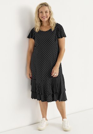 Smilende kvinde med blondt hår iført en sort kjole med hvide prikker og hvide sneakers, stående foran en ensfarvet hvid væg.