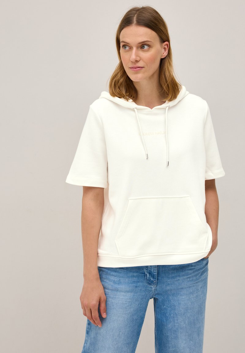 Witte hoody met een voorzak, korte mouwen, koord en een klein goud geborduurd logo op de borst. Gecombineerd met lichtblauwe jeans.