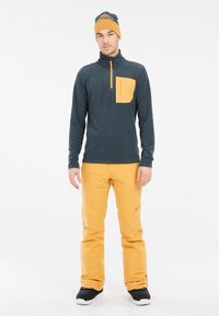 Fleece pullover in donkerblauw met een contrasterende gele borstzak; gecombineerd met gele broek en zwarte schoenen. Eenvoudig, functioneel ontwerp.