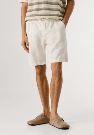 Hombre con zapatos sin cordones beige, pantalones cortos blancos con cordón y una camisa beige a rayas, de pie con una mano en el bolsillo.