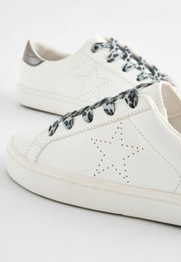 Białe skórzane sneakersy z perforowanym wzorem gwiazdy, czarno-szare sznurowadła w wzory oraz srebrny akcent na pięcie. Środkowa podeszwa jest lekko teksturowana.