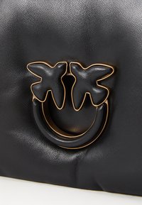 Bolsa de mão em couro preto com um acabamento texturizado. Apresenta dois detalhes em forma de borboleta e uma pega circular dourada ao centro.