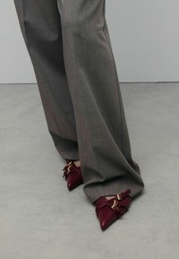 Chaussures bourgogne à bout pointu avec des sangles entrecroisées et des accents dorés, assorties à un pantalon gris ample en tissu texturé.