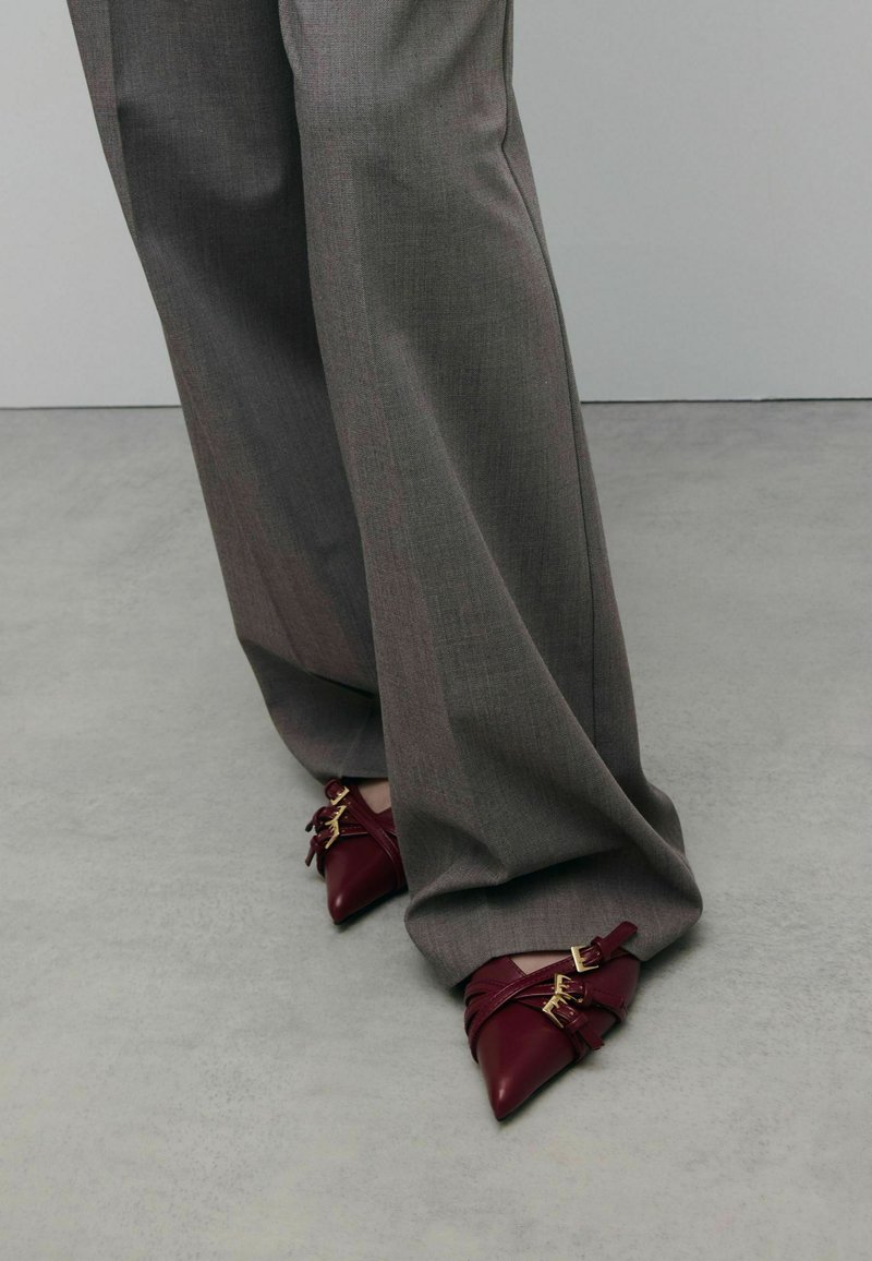 Chaussures bourgogne à bout pointu avec des sangles entrecroisées et des accents dorés, assorties à un pantalon gris ample en tissu texturé.