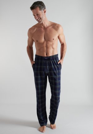 Oberkörperfreier Mann, der lächelt, barfuß steht mit den Händen in den Taschen, dunkle blaue karierte Pyjamahosen trägt vor schlichtem Hintergrund.