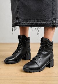 Black leather ankle boots with a block heel, featuring metal eyelets, a lace-up design, and a padded collar. Textured sole for grip.