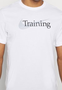 Camiseta de algodón blanca con un estampado negro de "Training" y un logo de Nike en azul claro sobre el texto. Mangas cortas, cuello redondo.