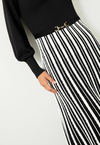 Melnas ribu virsotnes ar garām piedurknēm, apvienota ar baltu svītrainu svārku. Svārkiem ir viļņota tekstūra un zelta akcenti jostasvietā.