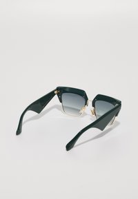 Gafas de sol de plástico verde con forma hexagonal, lentes en degradado gris, acentos dorados en las bisagras y brazos anchos y aerodinámicos.