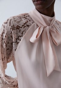 Blouse rose pâle avec manches longues en dentelle et détail de col noué. Caractérisée par une dentelle florale délicate et une texture de tissu lisse.