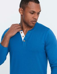 Man som bär en blå långärmad henley-tröja med tre träknappar, tittar ner och justerar kragen med sin vänstra hand.