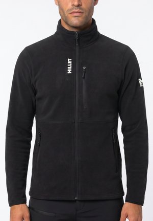 Mann trägt eine schwarze Fleecejacke mit Reißverschluss und vertikalem "MILLET"-Logo auf der Brust sowie einem Logo am linken Ärmel, steht vor weißem Hintergrund.