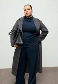 Palton gri de lână peste un pulover albastru închis cu guler înalt și pantaloni asortați. Geantă neagră cu design structurat. Texturi netede și fundal neutru.