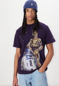 Lilla bomulds T-shirt med grafik af C-3PO og R2-D2. Modellen bærer en blå ribbet hue. Lyseblå jeans er synlige.