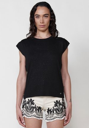 Camisetta nera a maniche corte con un sottile effetto glitter, abbinata a pantaloni corti beige con ricami floreali neri lungo l'orlo.
