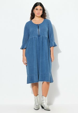 Denim-mekko keskipyörässä sinisessä, jossa on pyöreä kaula-aukko, puolihihat ja röyhelöhihoja. Siinä on edessä vetoketju ja liehuva helma.