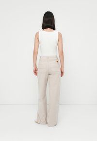 Donna che sta in piedi di spalle, con una maglietta bianca senza maniche, pantaloni beige a gamba larga e scarpe bianche su uno sfondo bianco uniforme.