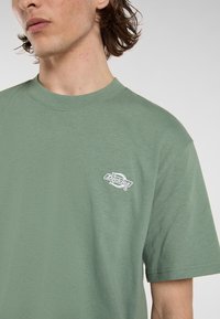 Grøn bomulds T-shirt med korte ærmer, rund halsudskæring og et lille hvidt Dickies-logo på venstre bryst. Blød tekstur, afslappet pasform.
