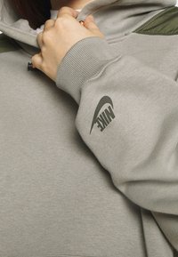 Sudadera de color gris claro con un acabado texturizado, que presenta un acento verde oscuro en el hombro y un logo negro de Nike en la manga.