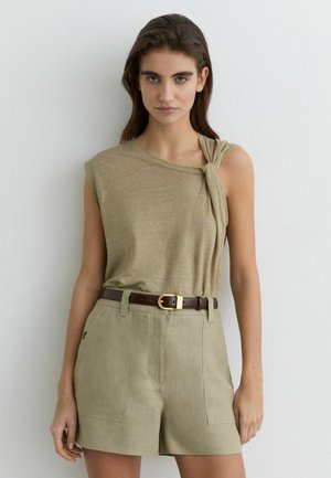 Kobieta o długich brązowych włosach, ubrana w beżowy bezrękawnik z węzłowym detalem oraz beżowe szorty z wysokim stanem i ciemnobrązowym paskiem.