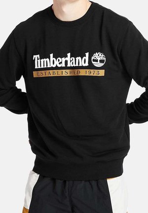 Osoba nosząca czarną bluzę Timberland z białym logo i napisem „Established 1973” na piersi.