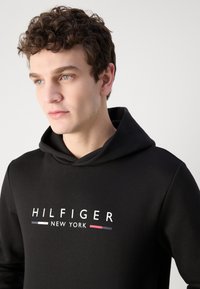 Sudadera negra hecha de un tejido suave y texturizado. Presenta una capucha con cordón y el logo "HILFIGER NEW YORK" en blanco, con acentos de color.