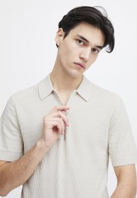 Beige kortermet polo-skjorte med teksturert strikkedesign og glidelås krage. Modell holder i glidelåsen med lysegrå detaljer.
