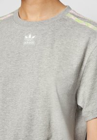 Grå bomull t-shirt med rund hals, korta ärmar och tre rosa och gröna ränder på axlarna. Adidas-logotyp på bröstet.