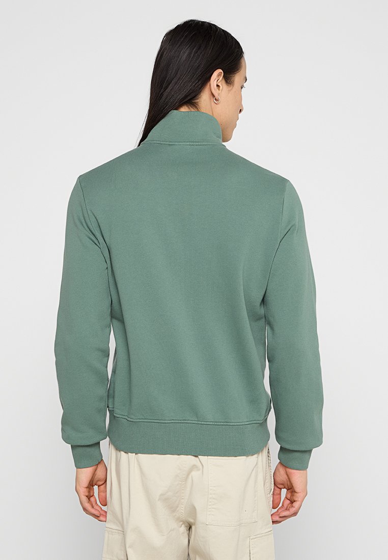 Persona con lunghi capelli scuri che indossa una felpa verde a colletto alto e pantaloni cargo beige, di spalle contro uno sfondo neutro.