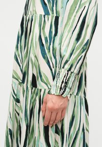 Χέρι που ακουμπά δίπλα σε φόρεμα με σχέδιο από φύλλα σε πράσινο, μπλε και μαύρο, με νερομπογιές, και μανσέτες με ραβδώσεις σε μακριά μανίκια.