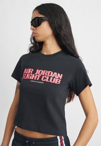 Νεαρή γυναίκα που φορά μαύρα γυαλιά ηλίου και μαύρο κοντομάνικο μπλουζάκι με την ένδειξη "Επίσημο Μέλος του Air Jordan Flight Club" σε ροζ κείμενο.