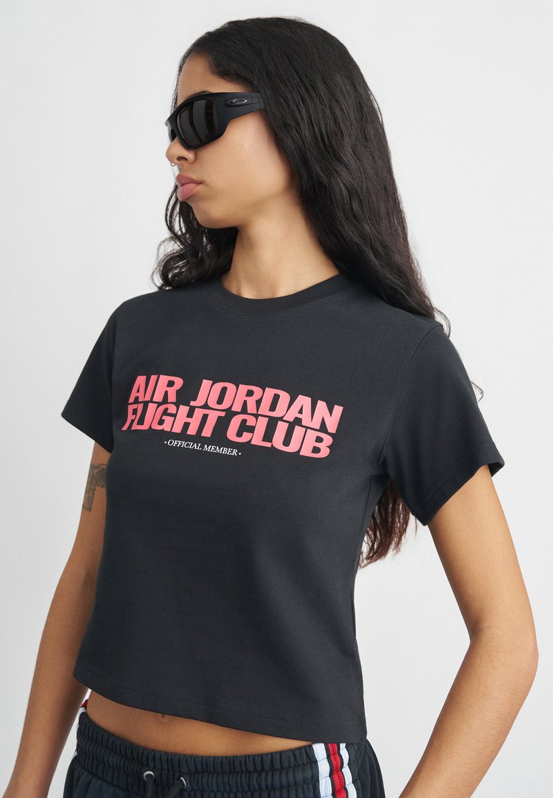 Νεαρή γυναίκα που φορά μαύρα γυαλιά ηλίου και μαύρο κοντομάνικο μπλουζάκι με την ένδειξη "Επίσημο Μέλος του Air Jordan Flight Club" σε ροζ κείμενο.