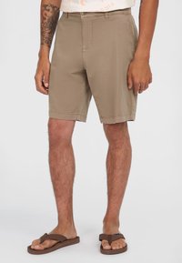 Beige Shorts mit geradem Schnitt, aus leichtem Stoff. Verfügt über Fronttaschen und einen Reißverschluss. Getragen mit braunen Flip-Flops.