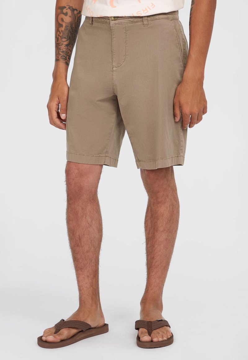 Beige Shorts mit geradem Schnitt, aus leichtem Stoff. Verfügt über Fronttaschen und einen Reißverschluss. Getragen mit braunen Flip-Flops.